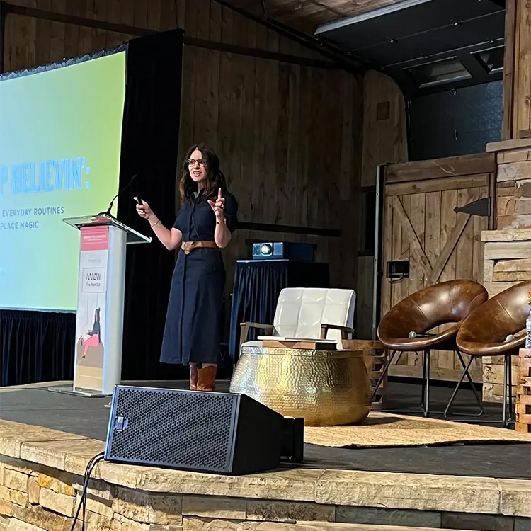 Female keynote speaker addressing an audience on a rustic stage beside a podium and display screen.
