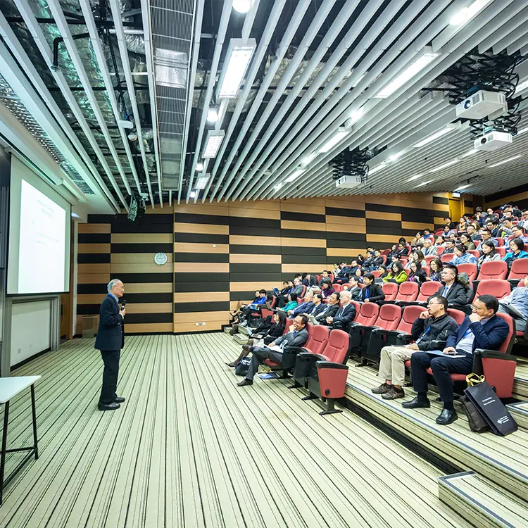 Speaker presenting with a microphone to a seated audience in a modern tiered lecture hall.